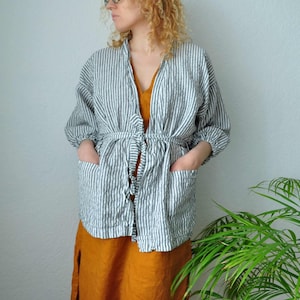 Puede incluir: Un kimono de lino a rayas con cintur&oacute;n de lazo y bolsillos. El kimono es blanco con finas rayas negras. La modelo lleva un vestido amarillo mostaza debajo del kimono.