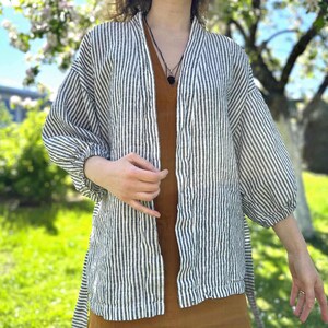 Puede incluir: Un kimono de lino a rayas blancas y negras con mangas largas y cierre de lazo. El kimono se lleva sobre un vestido marr&oacute;n.