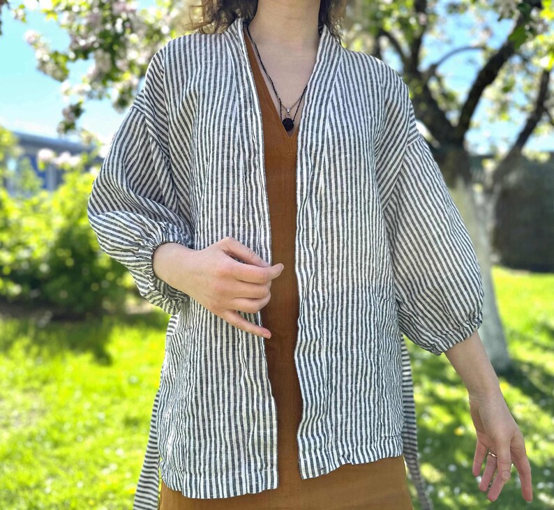 Puede incluir: Un kimono de lino a rayas blancas y negras con mangas largas y cierre de lazo. El kimono se lleva sobre un vestido marr&oacute;n.