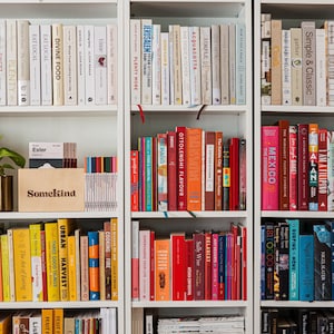 Könnte beinhalten: Ein weißes Bücherregal mit einer Vielzahl von Büchern. Die Bücher sind nach Farben geordnet, mit Buchrücken in Weiß-, Gelb-, Orange-, Rot- und Blautönen. Eine kleine Pflanze in einem goldenen Topf und eine Box mit dem Wort "Somekind" sind ebenfalls zu sehen.