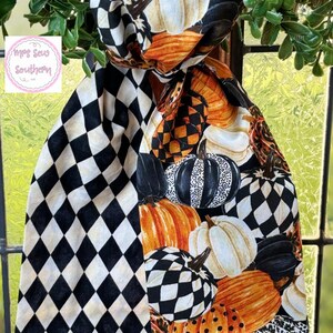 Op de afbeelding: Een decoratieve herfst sjaal met een patroon van pompoenen in oranje, wit, zwart en geblokte ontwerpen. De sjaal is over een raam gedrapeerd, met groene bladeren op de achtergrond. De sjaal heeft een zwart-wit ruitpatroon.