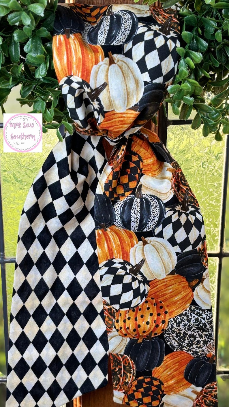 Op de afbeelding: Een decoratieve herfst sjaal met een patroon van pompoenen in oranje, wit, zwart en geblokte ontwerpen. De sjaal is over een raam gedrapeerd, met groene bladeren op de achtergrond. De sjaal heeft een zwart-wit ruitpatroon.