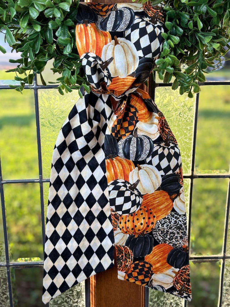 Op de afbeelding: Decoratieve sjaal met herfstthema met een mix van pompoenpatronen en een zwart-wit ruitpatroon. De pompoenen zijn in tinten oranje, wit en zwart. De sjaal is gedrapeerd over een raamkozijn met een groene bladkrans.