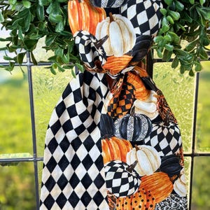 Op de afbeelding: Decoratieve sjaal met herfstthema met een patroon van pompoenen in verschillende kleuren, waaronder oranje, wit en zwart. De sjaal heeft ook een zwart-wit ruitpatroon. De sjaal is gedrapeerd over een raam met groen gebladerte.