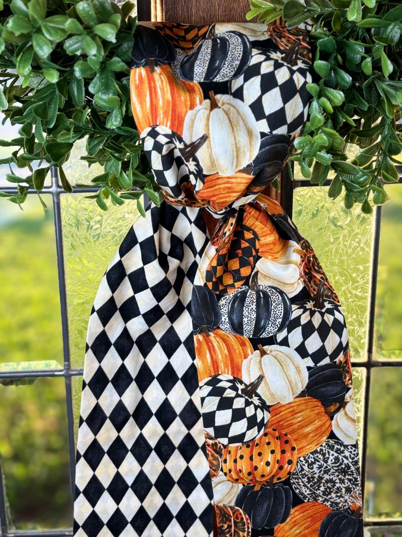 Op de afbeelding: Decoratieve sjaal met herfstthema met een patroon van pompoenen in verschillende kleuren, waaronder oranje, wit en zwart. De sjaal heeft ook een zwart-wit ruitpatroon. De sjaal is gedrapeerd over een raam met groen gebladerte.