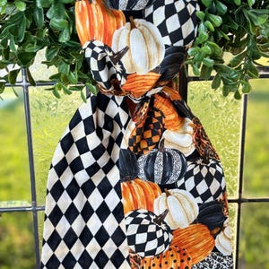 Op de afbeelding: Een decoratieve sjaal met een herfstthema, met pompoenen in oranje, wit en zwart. De sjaal heeft een zwart-wit ruitpatroon en wordt omlijst door groene bladeren. De pompoenen hebben verschillende patronen en texturen.