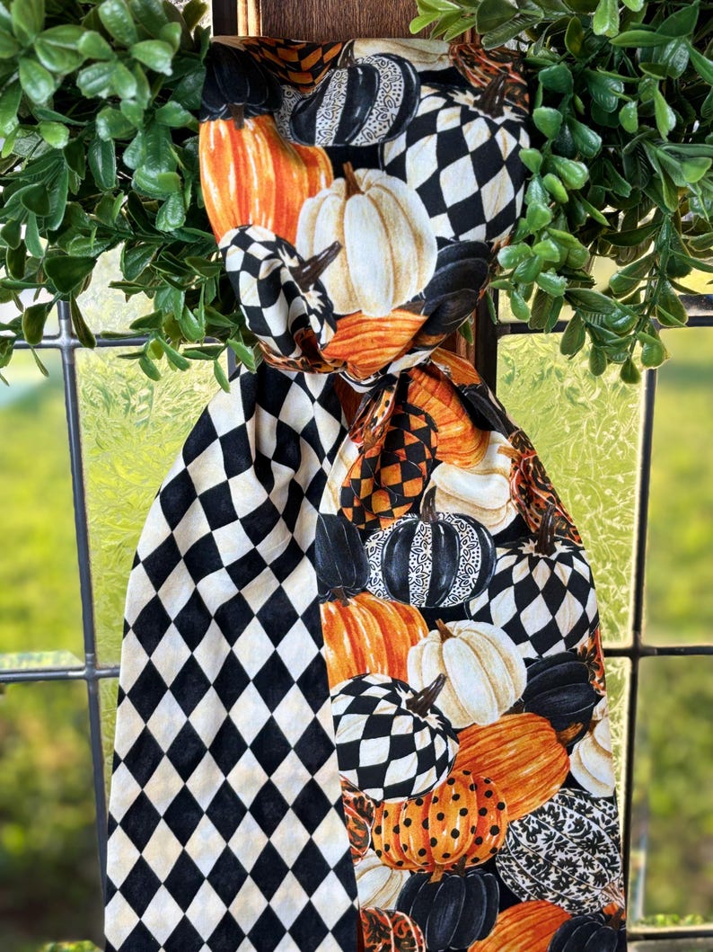 Op de afbeelding: Een decoratieve sjaal met een herfstthema, met pompoenen in oranje, wit en zwart. De sjaal heeft een zwart-wit ruitpatroon en wordt omlijst door groene bladeren. De pompoenen hebben verschillende patronen en texturen.