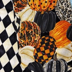 Op de afbeelding: Een stof met een herhalend patroon van pompoenen in verschillende kleuren en ontwerpen, waaronder oranje met zwarte stippen, zwart en cr&egrave;me. De pompoenen staan tegen een zwarte achtergrond, met een zwart-wit ruitpatroon aan de linkerkant.