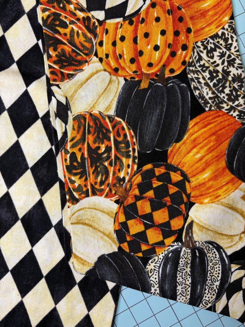 Op de afbeelding: Een stof met een herhalend patroon van pompoenen in verschillende kleuren en ontwerpen, waaronder oranje met zwarte stippen, zwart en cr&egrave;me. De pompoenen staan tegen een zwarte achtergrond, met een zwart-wit ruitpatroon aan de linkerkant.