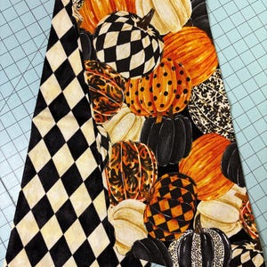 Op de afbeelding: Een decoratieve stof met een herhalend patroon van pompoenen in verschillende kleuren en patronen, waaronder oranje, wit, zwart en geblokte ontwerpen. De stof heeft ook een zwart-wit ruitpatroon.