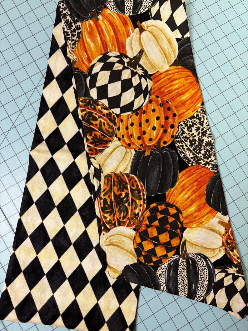 Op de afbeelding: Een decoratieve stof met een herhalend patroon van pompoenen in verschillende kleuren en patronen, waaronder oranje, wit, zwart en geblokte ontwerpen. De stof heeft ook een zwart-wit ruitpatroon.