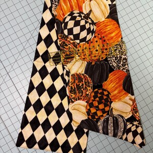 Op de afbeelding: Een stuk stof met een patroon van pompoenen in oranje, wit, zwart en dambordpatronen. De linkerkant heeft een zwart-wit ruitpatroon. Geschikt voor herfst- of Halloween-knutselwerk.