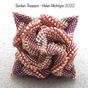 May include: A beaded rose-shaped sculpture in shades of pink, copper, and gold. The sculpture is made of small, iridescent beads and has a complex, intricate design. "Sunken Treasure - Helen McIntyre 2022" is written above the sculpture.