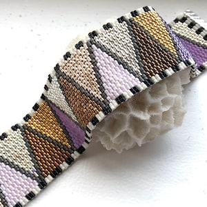 May include: A beaded bracelet with a geometric pattern in shades of white, black, gold, and purple.