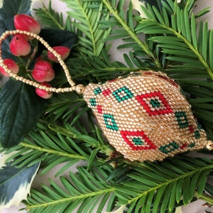 May include: A gold beaded Christmas bauble with a hanging loop. The bauble features a pattern of red and green diamond shapes. It rests on green pine needles and red berries, creating a festive display.