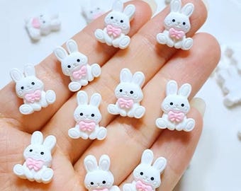 May include: A collection of white bunny-shaped embellishments with pink bow ties. The bunnies have black eyes and are sitting down. These small, decorative bunnies are ideal for crafts and DIY projects.