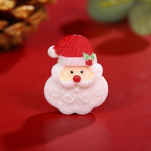 May include: A small, white, glittery Santa Claus figurine with a red hat and a green leaf on top. The figurine has a red nose and black eyes.