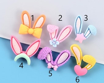 May include: Six different pastel colored bunny ear charms with various embellishments. The charms are numbered 1 through 6. Charm 1 has a yellow bow, charm 2 has a blue flower, charm 3 has a pink star, charm 4 has a green headband, charm 5 has a purple bow, and charm 6 has a pink heart.