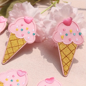 Puede incluir: Parches bordados con forma de conos de helado. El helado rosa tiene un corazón y chispas de colores. El cono es amarillo dorado con un patrón de entramado. Los parches están sobre un fondo rosa.