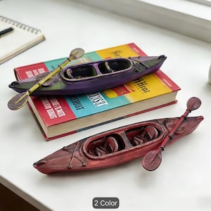 Op de afbeelding: Twee miniatuur kajakbeeldjes, een paarse en een rode, elk met een dubbelbladige peddel die bovenop rust. De kajaks hebben gedetailleerde cockpits met gevormde stoeltjes en gestructureerde rompen. De tekst 2 Color is onderaan zichtbaar.