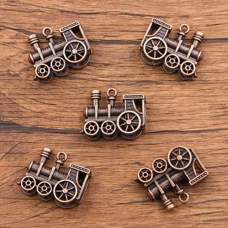 May include: Five antique-style metal train engine charms with intricate details. Each charm features a locomotive with wheels, a smokestack, and a bell. The charms are a dark bronze color.