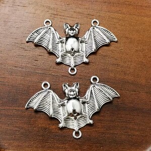 May include: Two silver-toned metal bat charms featuring detailed wing structures and a central body. Each charm includes three small circular loops positioned at the top of each wing and at the bottom of the bat for attachment purposes.