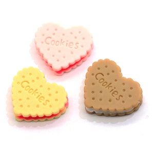 Könnte beinhalten: Drei herzförmige Kekse mit rosa, gelbem und braunem Zuckerguss. Jeder Keks trägt die Aufschrift "Cookies".
