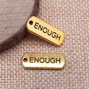 May include: Two gold-coloured metal tags with the word "ENOUGH" engraved on them. The tags have a hole at the top for attaching to a chain or string.