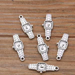 Może przedstawiać: Srebrne zawieszki do zegarków z prostokątną tarczą i połączonym paskiem. Tarcza zegarka ma szczegółowy wzór zegara. Każdy urok ma małą pętlę u góry i u dołu do mocowania. Ozdoby są rozrzucone na drewnianej powierzchni.