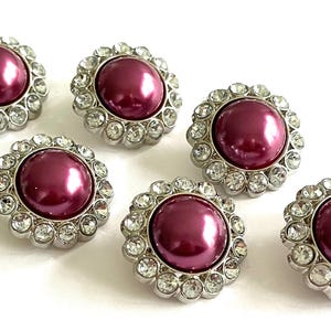 May include: Six decorative buttons with a central, round, deep pink pearl surrounded by a ring of clear, sparkling rhinestones. The buttons have a silver-colored metal base and are arranged on a white background.