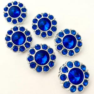 May include: Six blue rhinestone buttons with a silver-colored metal rim. Each button features a large, faceted center stone surrounded by smaller, round rhinestones. The buttons are arranged on a white background.