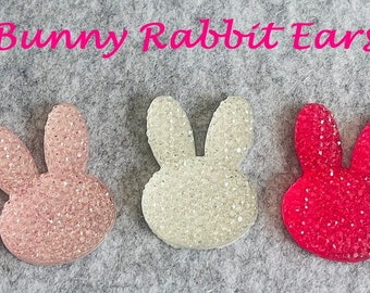 May include: Three sparkly bunny rabbit ears in pink, white, and red. The ears are made of a glittery material and are shaped like bunny ears.