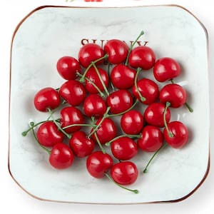 Puede incluir: Un plato cuadrado blanco con un diseño de mármol y borde dorado, lleno de cerezas rojas brillantes con tallos verdes. El plato tiene la palabra "SUNNY" impresa.