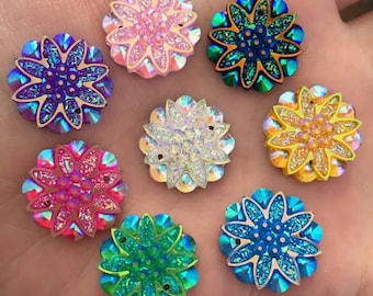 May include: A collection of eight flower-shaped embellishments with iridescent, glittery petals. The color palette includes pink, purple, blue, green, white, and yellow. Each flower has a central, sparkling design, and a metallic border.