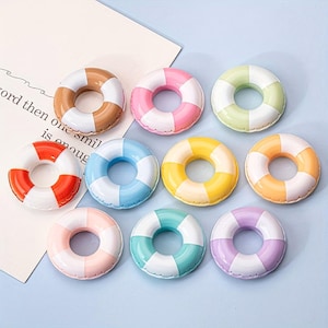 Könnte beinhalten: Neun farbenfrohe Miniatur-Rettungsringe in verschiedenen Farben, darunter Braun, Rosa, Grün, Rot, Blau, Gelb, Orange, Türkis und Lila. Jeder Ring hat ein weißes Streifendesign. Die Objekte sind auf einer hellblauen Oberfläche angeordnet.