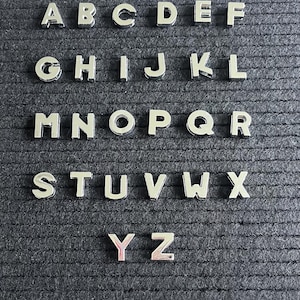 Puede incluir: Un conjunto de fichas de letras plateadas, dispuestas en orden alfab&eacute;tico, sobre un fondo negro texturizado. Las letras son de la A a la Z.