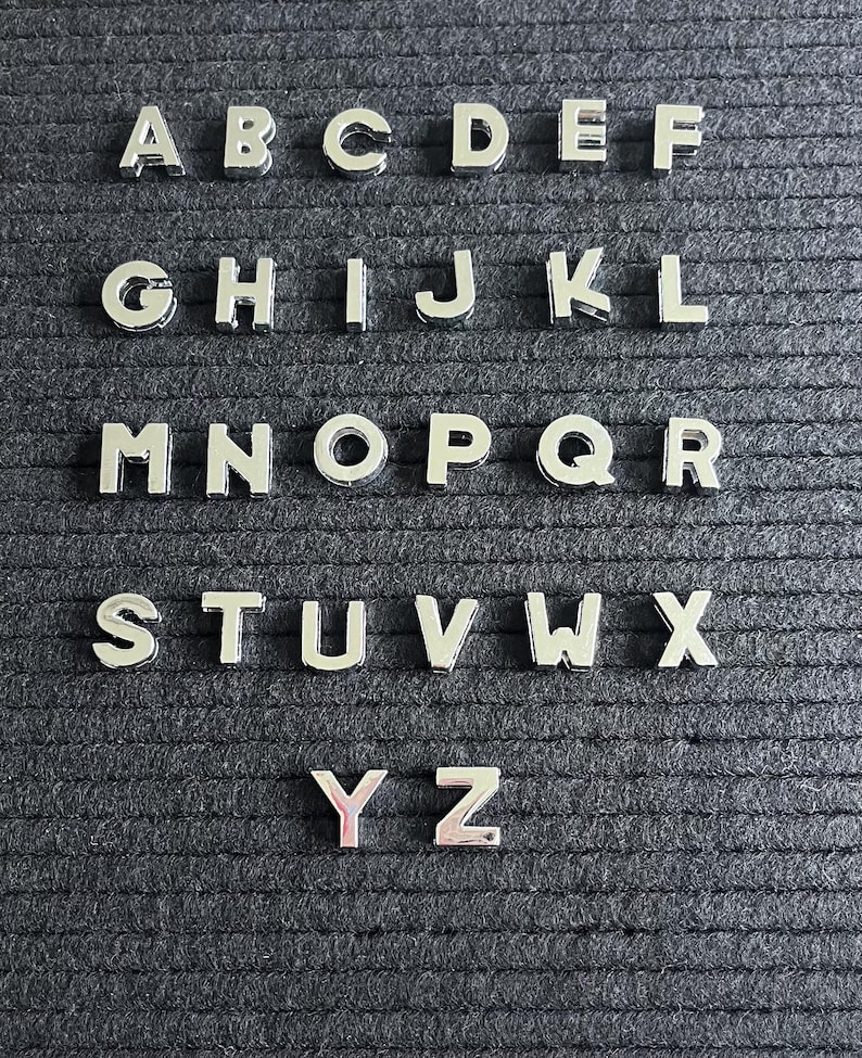 Puede incluir: Un conjunto de fichas de letras plateadas, dispuestas en orden alfab&eacute;tico, sobre un fondo negro texturizado. Las letras son de la A a la Z.