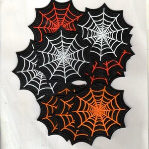 May include: A collection of embroidered patches shaped like spider webs. The patches feature black fabric backgrounds with stitched web designs in contrasting colors including bright orange, vibrant red, and stark white thread.