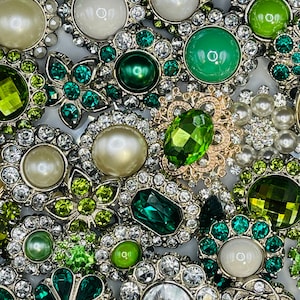 May include: Assortment of green and clear rhinestone embellishments with silver settings. The embellishments feature various shapes, including flowers, hearts, and geometric designs.