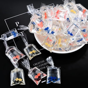 Puede incluir: Una colección de pequeñas bolsas de plástico llenas de peces de plástico de colores. Las bolsas están atadas con cuerda y están dispuestas en círculo sobre un plato blanco. El plato está sobre una superficie negra.