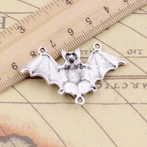 May include: A silver-toned metal bat charm with three small circular loops for attachment. The charm is positioned next to a wooden ruler showing a width of approximately 4.5 cm from wingtip to wingtip, with detailed textured wings.