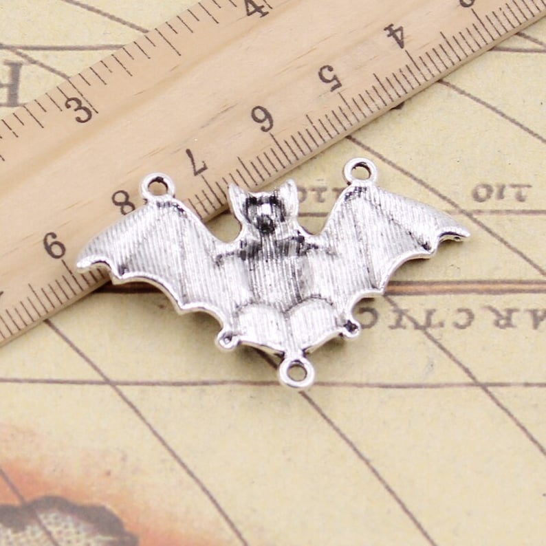 May include: A silver-toned metal bat charm with three small circular loops for attachment. The charm is positioned next to a wooden ruler showing a width of approximately 4.5 cm from wingtip to wingtip, with detailed textured wings.