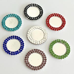 May include: A collection of decorative, round coasters with a mirrored center and a border of colorful rhinestones. Colors include teal, black, red, silver, blue, purple, and green. These coasters are designed to protect surfaces from drink rings.