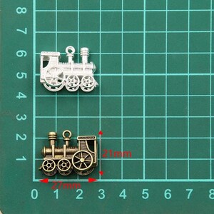 May include: Two silver and bronze train charms, one is 21 mm long and the other is 27 mm long. The charms are on a green grid background.