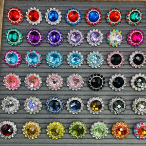 May include: Assortment of 36 colorful rhinestone embellishments with a variety of colors and designs. Each embellishment features a round, faceted rhinestone surrounded by a silver-toned metal frame.
