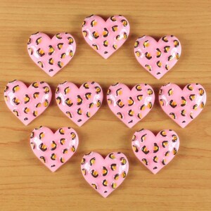 May include: Ten pink heart-shaped decorative objects with a leopard print pattern in yellow and black. The hearts are arranged on a light brown wooden surface. The objects have a glossy finish.