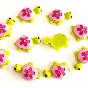 Puede incluir: Colección de pequeñas figuras decorativas de tortugas. Cada tortuga tiene un cuerpo amarillo, un caparazón rosa con una flor rosa oscuro y ojos negros. Las tortugas están dispuestas en un patrón circular.