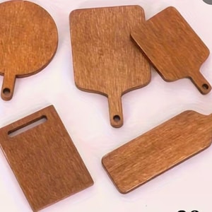 Puede incluir: Un conjunto de tablas de cortar de madera de varias formas. El conjunto incluye una tabla redonda con asa, una tabla rectangular con asa, dos tablas en forma de paleta y una tabla rectangular con un recorte para el asa. Las tablas tienen una veta de madera cálida y natural.