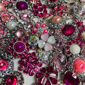 May include: Assortment of pink and clear rhinestone embellishments in various shapes, including hearts, flowers, and geometric designs. The embellishments are set in silver metal.