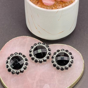 May include: Three black gemstone buttons with a silver-colored border are displayed on a pink, gold-edged surface. The buttons have a faceted center and are surrounded by smaller black stones. A white pot with a plant is in the background.